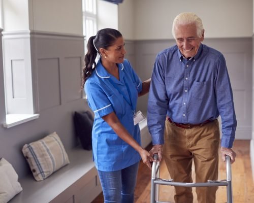 Senior Man At Home Using Walking Frame Being Helped By Female Ca Senior Man At Home Using Walking Frame Being Helped By Female Care Worker In Uniform